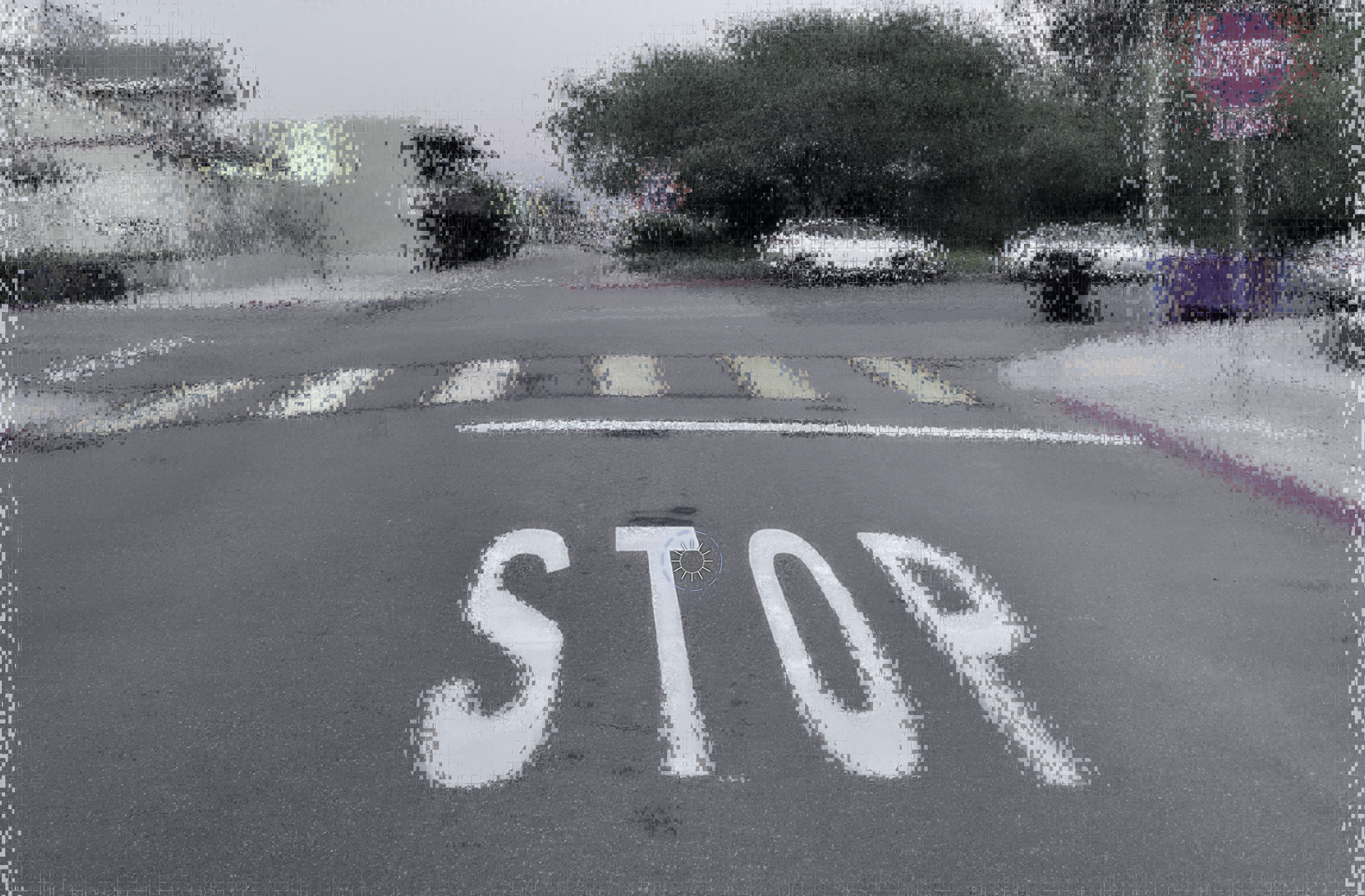 Street scene through Scrutinizer's peripheral filter, fixated on the white STOP painted on the road: the foveal region holds detail; the red octagonal stop sign in the upper-right corner desaturates and loses chromatic identity at peripheral eccentricity